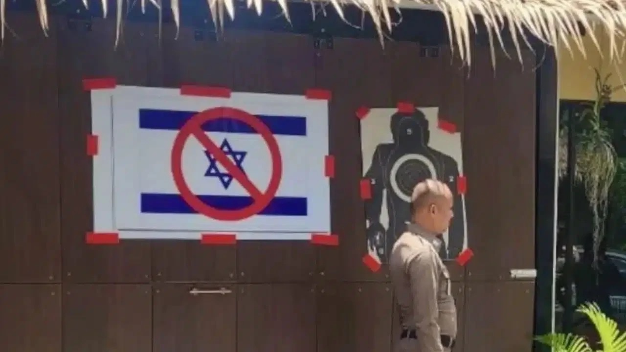 En uppsatt skylt med en överkryssad israelisk flagga bredvid en skottavla på en vägg vid djurparken, med en thailändsk polis framför.